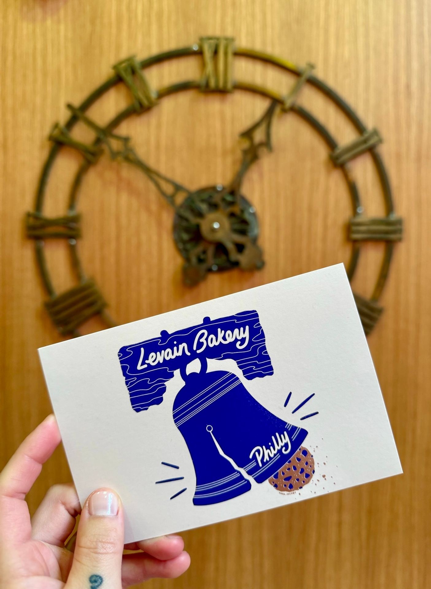 Hand holding a Levain Bakery Philly card in front of a large decorative wall clock.